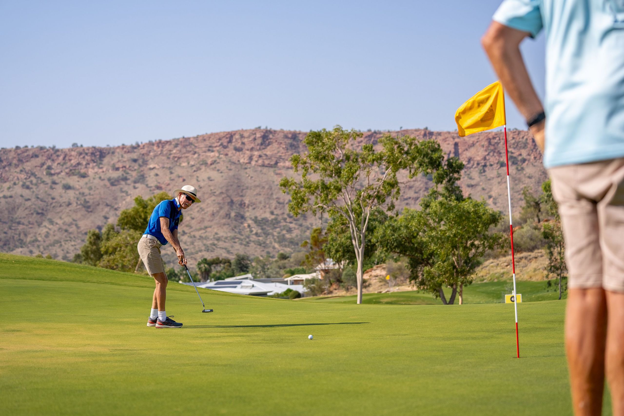 A golfer in a blue shirt and shorts putts on a green, with a yellow flag nearby and rocky hills in the background.