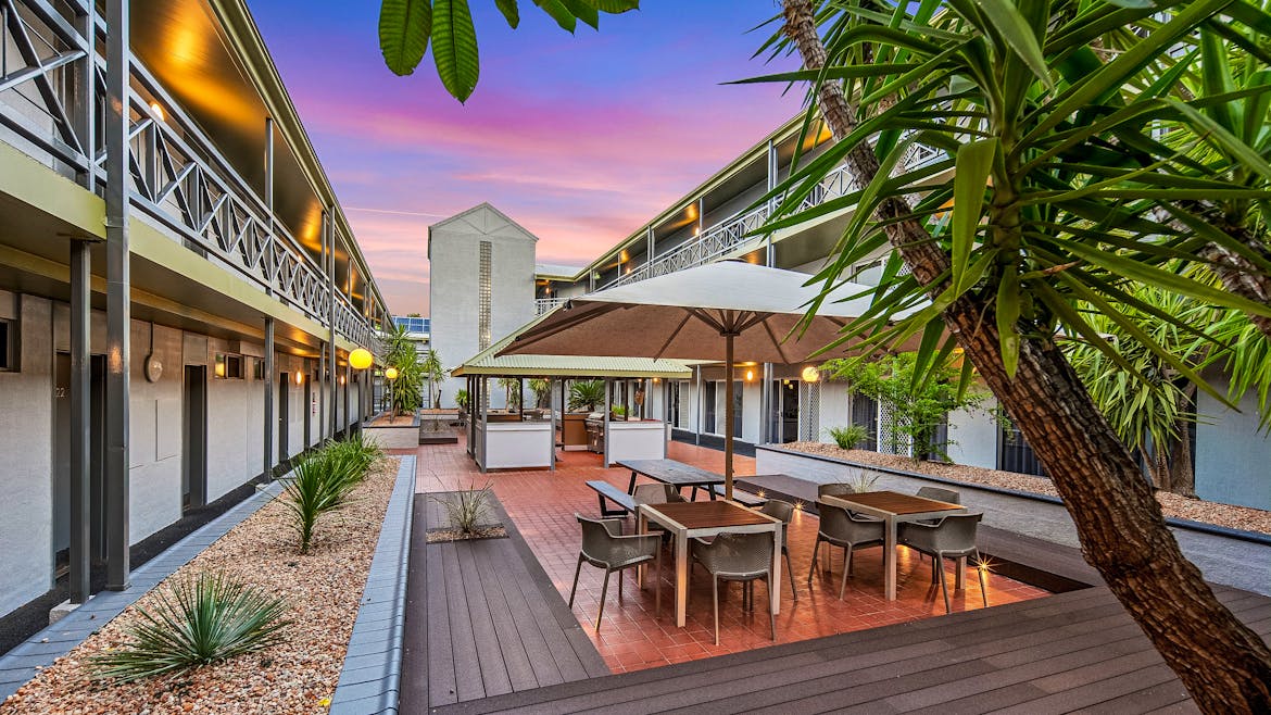 Outdoor courtyard with tables and chairs, surrounded by modern two-story buildings. The sky is vibrant with pink and purple hues at sunset.