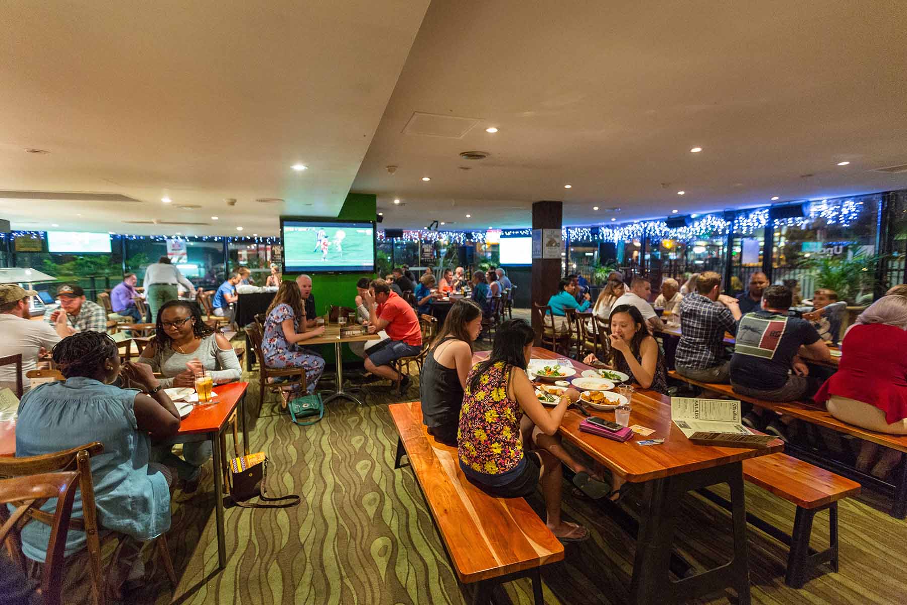 A lively restaurant scene with people dining at wooden tables, watching sports on TV, and enjoying a festive atmosphere with string lights.