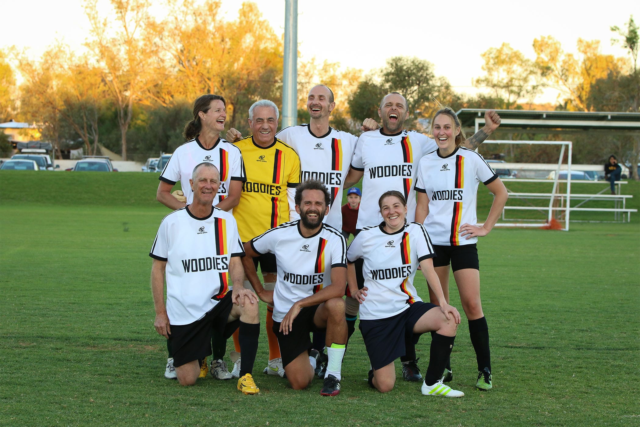A smiling co-ed soccer team, the "Woodies," poses for a group photo on a grassy field in their matching white, black, and yellow uniforms.