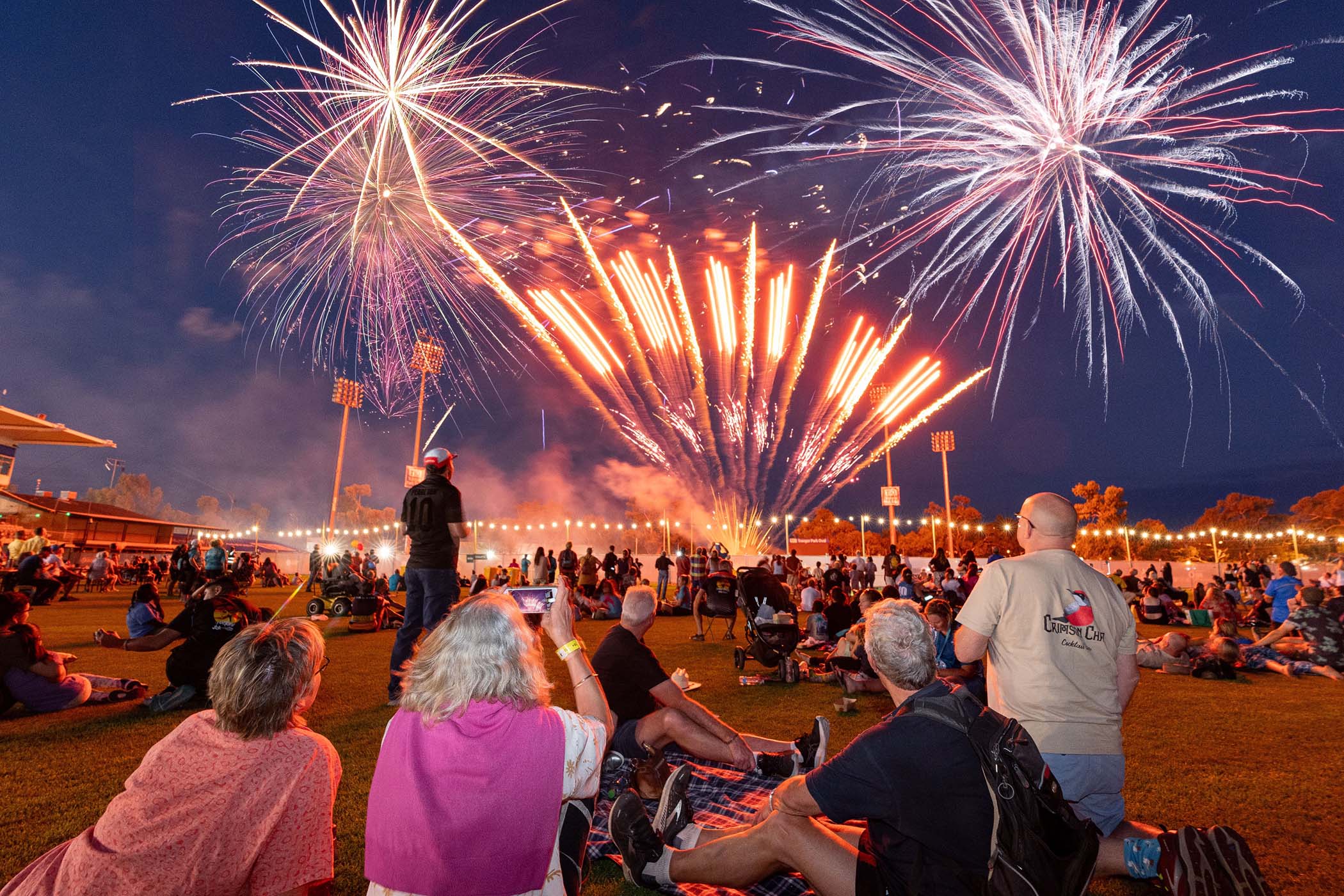 A crowd of people sits on a grassy field at night, watching a spectacular display of red and white fireworks light up the dark sky.
