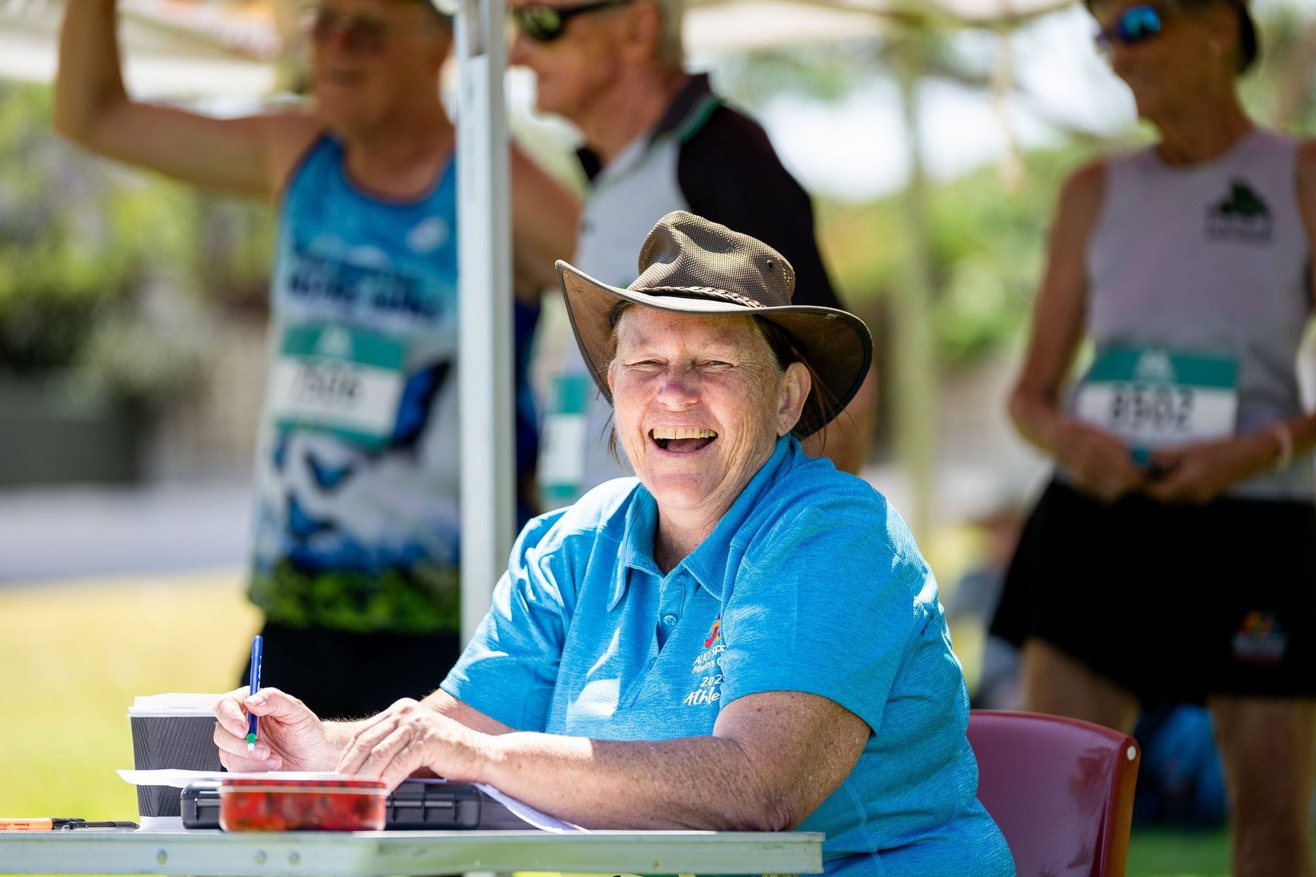 A smiling woman in a hat sits at a table outdoors, writing. People in athletic wear are in the background.