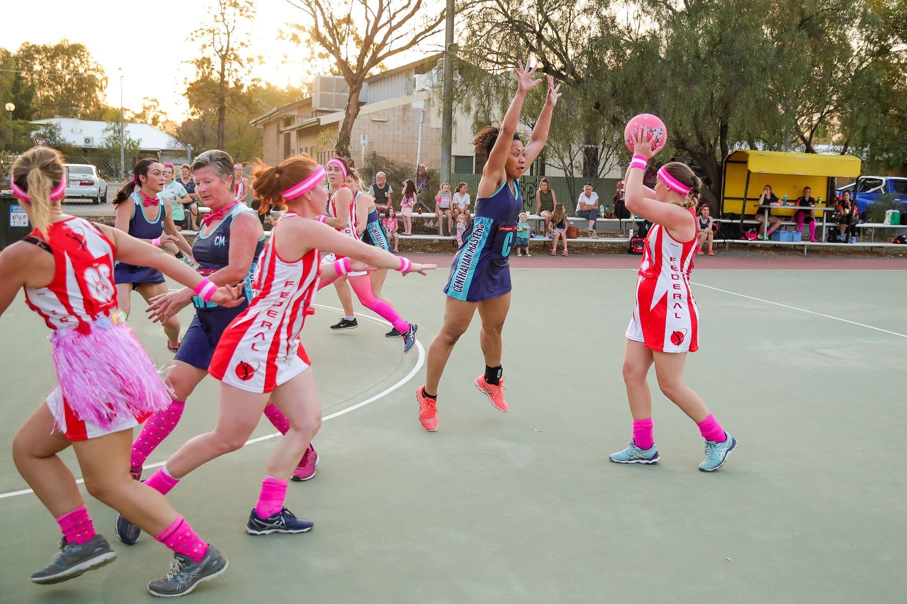 Women playing netball on an outdoor court, with one player jumping to block a pass. All wear colorful uniforms and pink socks. Spectators watch nearby.