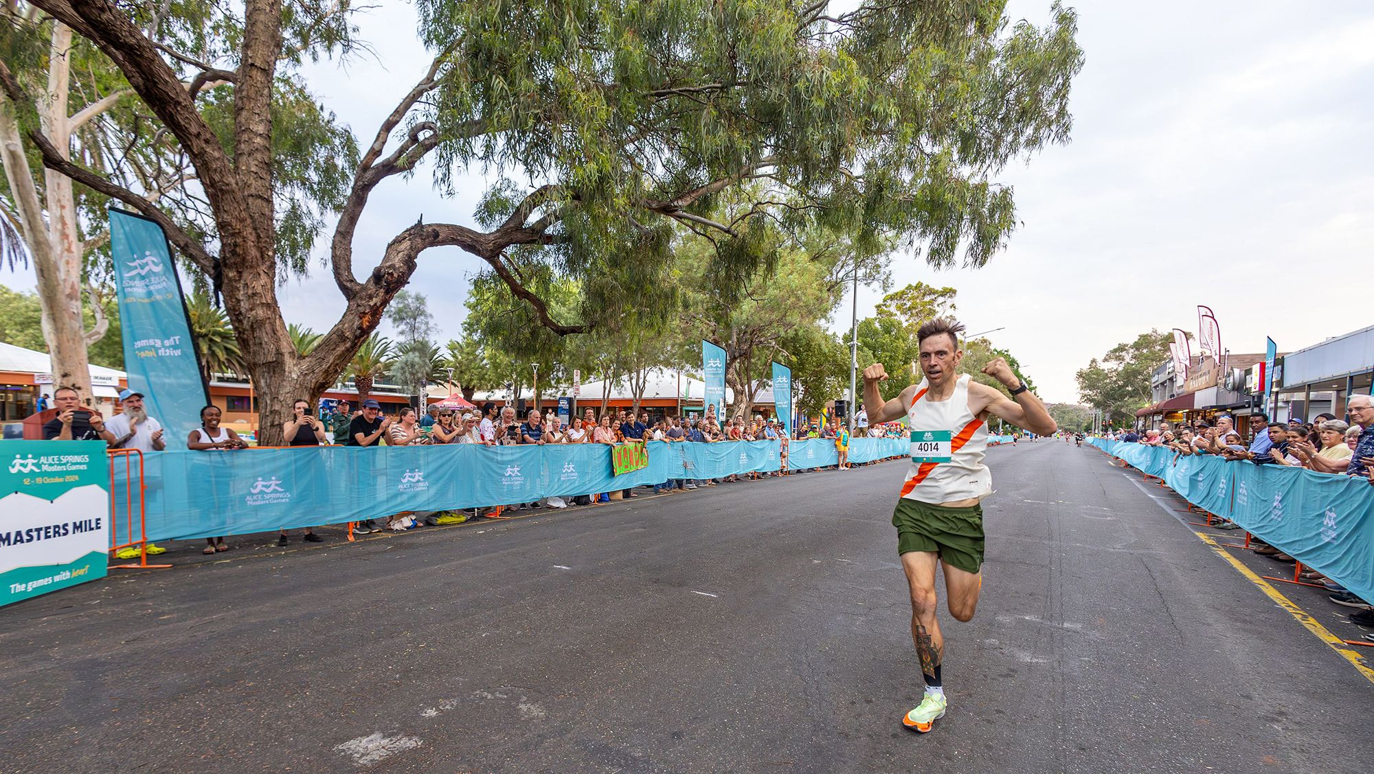 A runner in athletic gear celebrates crossing the finish line of a street race, surrounded by cheering spectators and event banners.