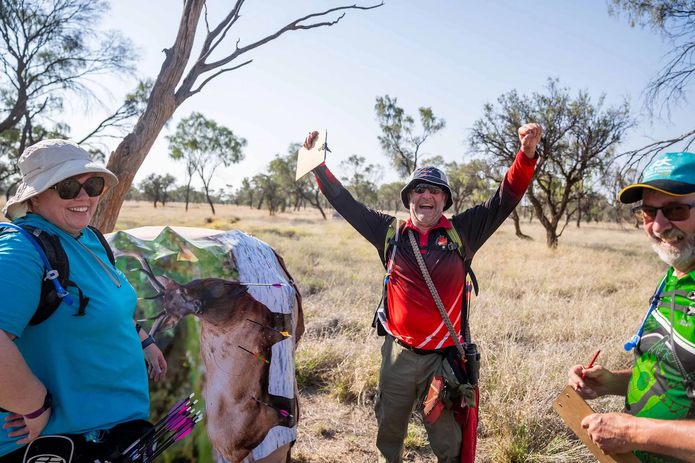 Three archers smile beside a wildlife target in the Australian bush, with the central competitor raising both arms in celebration.