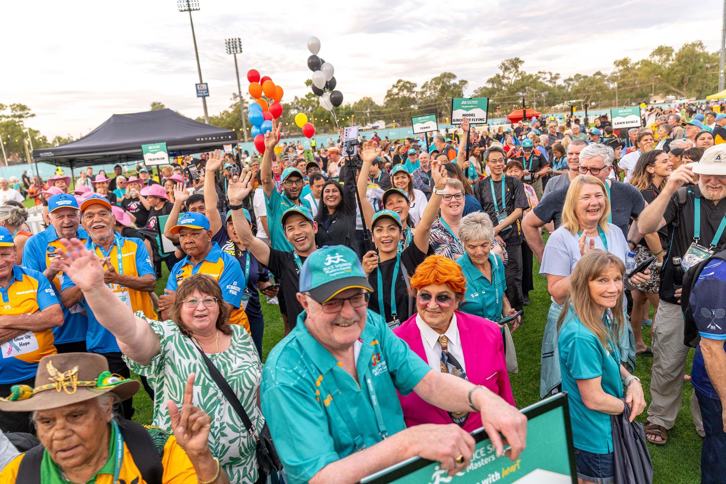 A large, diverse crowd of cheerful participants waves and smiles at an outdoor sporting event, with balloons and signs visible in the background.