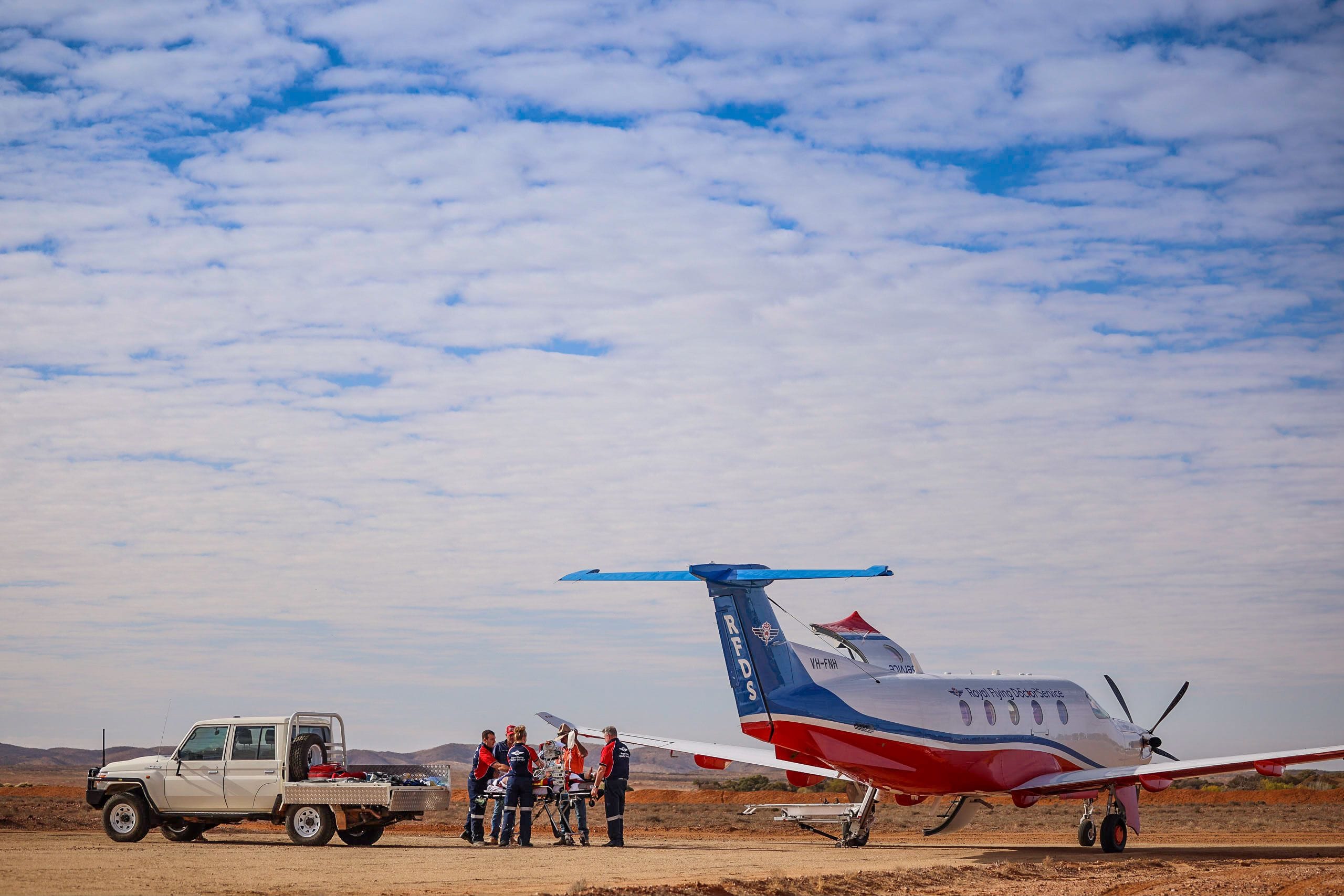 A Royal Flying Doctor Service team transfers a patient on a stretcher from a truck to their plane on a remote dirt airstrip under a vast, cloudy sky.