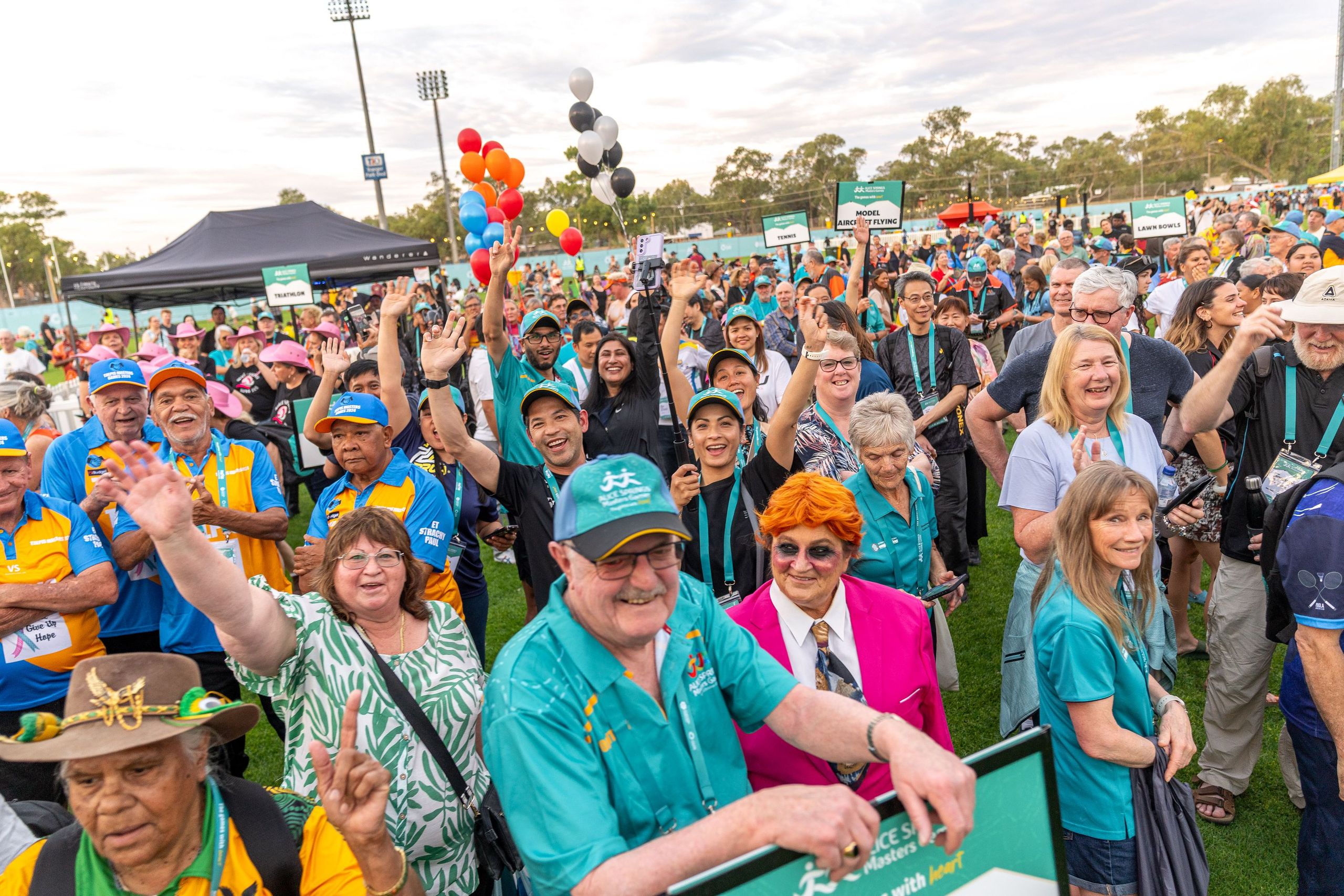 A diverse group of people in colorful attire cheer and wave at an outdoor event, surrounded by balloons and tents.